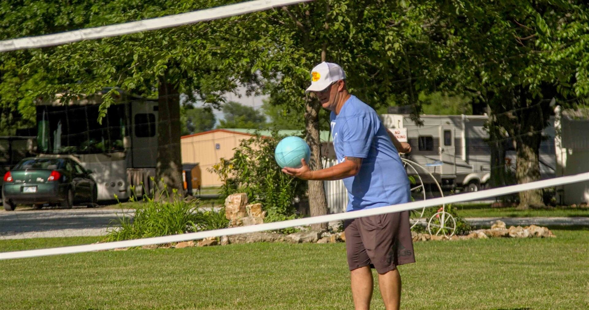 A person holding a blue ball in a grassy area with trees and a fence in the background.
