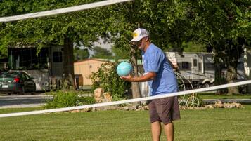 A person holding a blue ball in a grassy area with trees and a fence in the background.