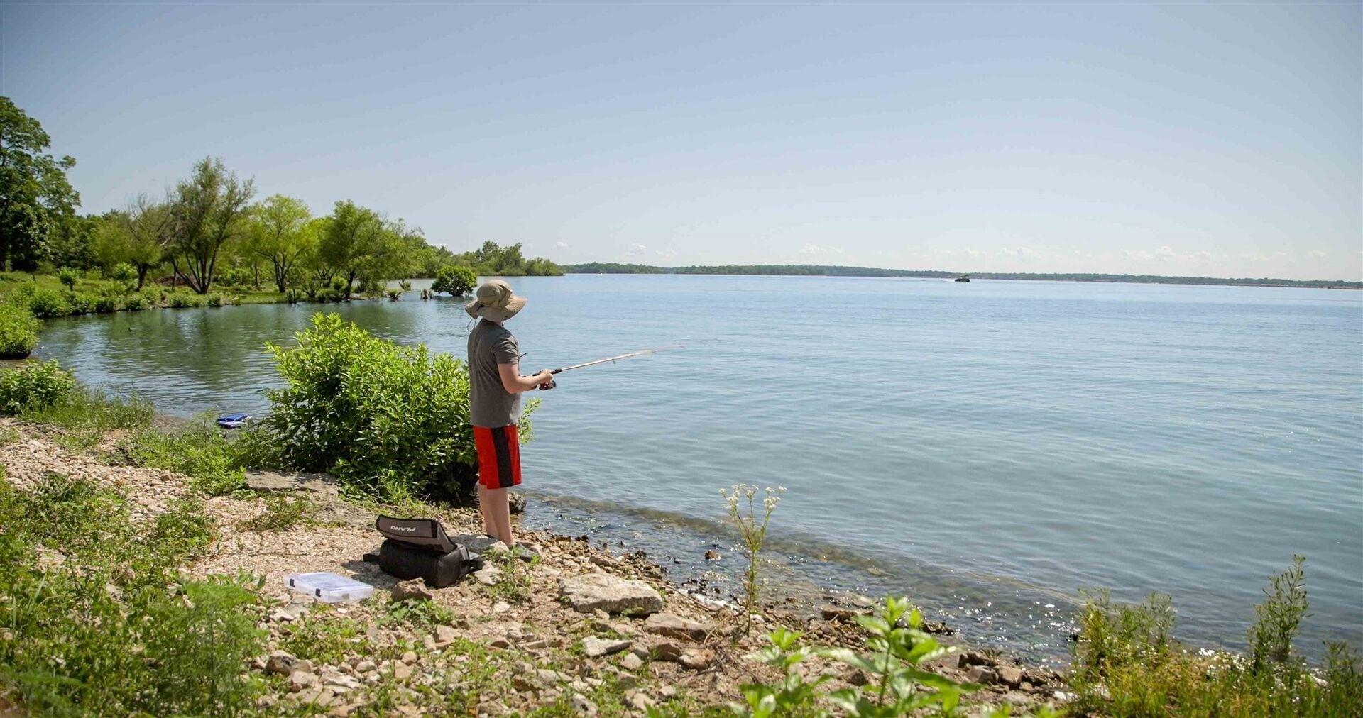A person fishing by a calm lake with trees in the background.