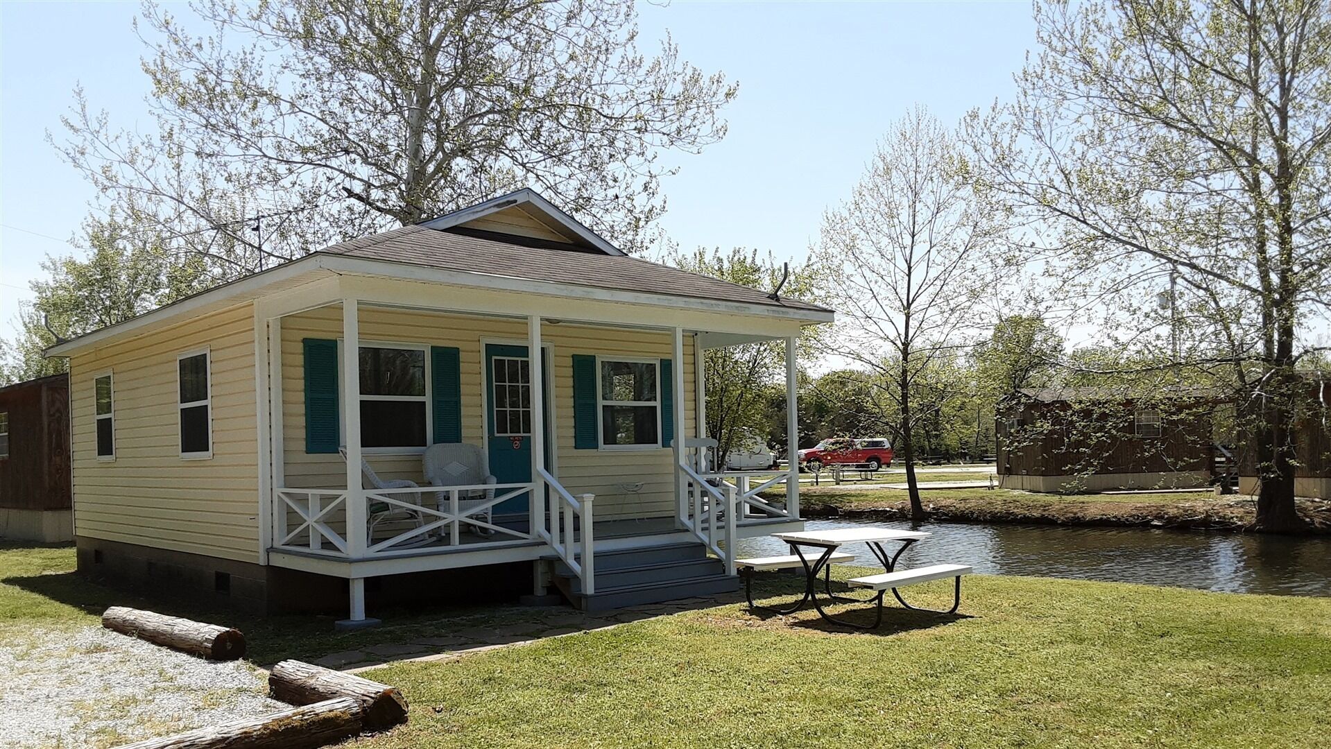 A small, single-story cabin with a porch, a picnic table, and a pond in the background.