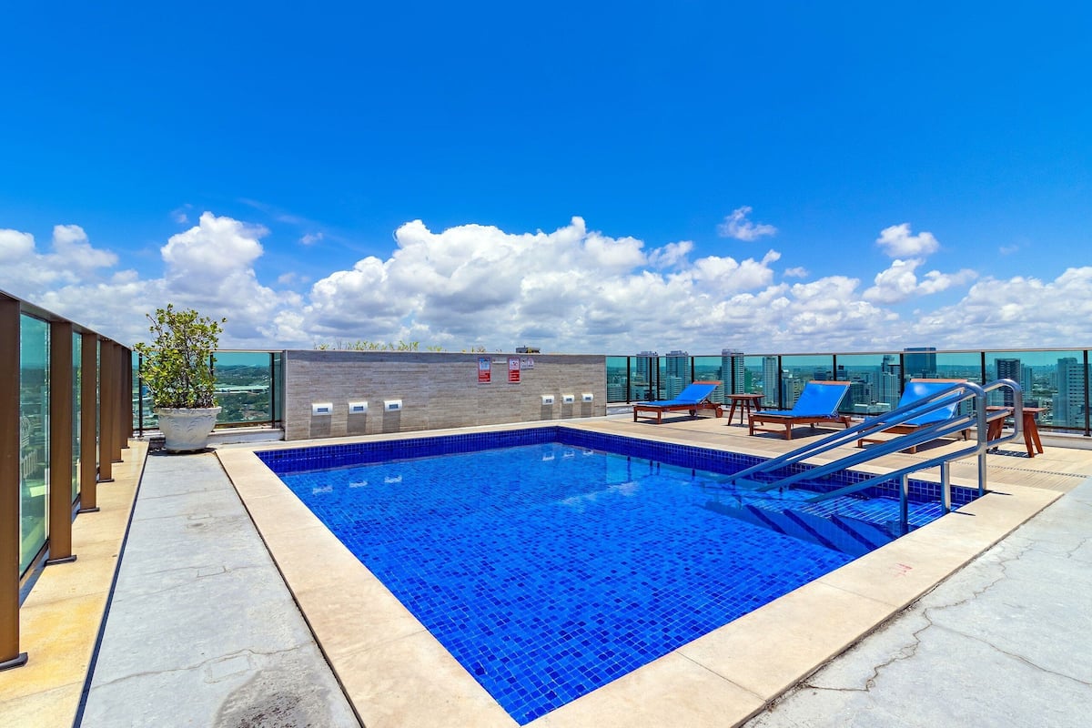 A rooftop pool with clear blue water, surrounded by lounge chairs and a potted plant.