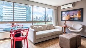 A modern living room with a sofa, ottoman, and red chairs, large windows with a city view, and a mounted air conditioner. - Home Stay #606 - Studio in Recife by Carpediem (Recife)