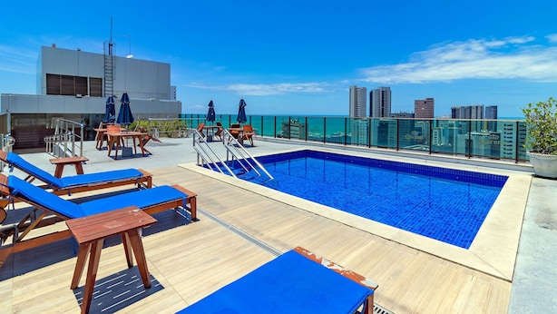 A rooftop pool with blue-tiled water, wooden lounge chairs, and a glass railing overlooking a cityscape. - Home Stay #606 - Studio in Recife by Carpediem (Recife)