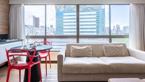 A modern living room with a sofa, red chairs, a dining table with cups, and a large window with a city view. - Home Stay #606 - Studio in Recife by Carpediem (Recife)