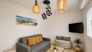En moderne stue med en grå sofa, en rund skammel, et lille bord og en potteplante. Der er to hængelamper og et fladskærms-tv monteret på væggen.