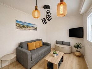 A modern living room with a grey sofa, a round ottoman, a small table, and a potted plant. There are two pendant lights and a flat-screen TV mounted on the wall. - Modern Semi-Basement Αpt - Vibrant Kypseli by ΜΡS (Athina)