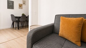 En moderne stue med en grå sofa, en pudebetræk i sennepsfarve, et spiseområde med stole og bord, samt et maritimt maleri på væggen.