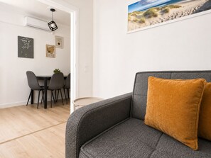 A modern living room with a grey sofa, a mustard-colored pillow, a dining area with chairs and a table, and a beach-themed painting on the wall. - Modern Semi-Basement Αpt - Vibrant Kypseli by ΜΡS (Athina)