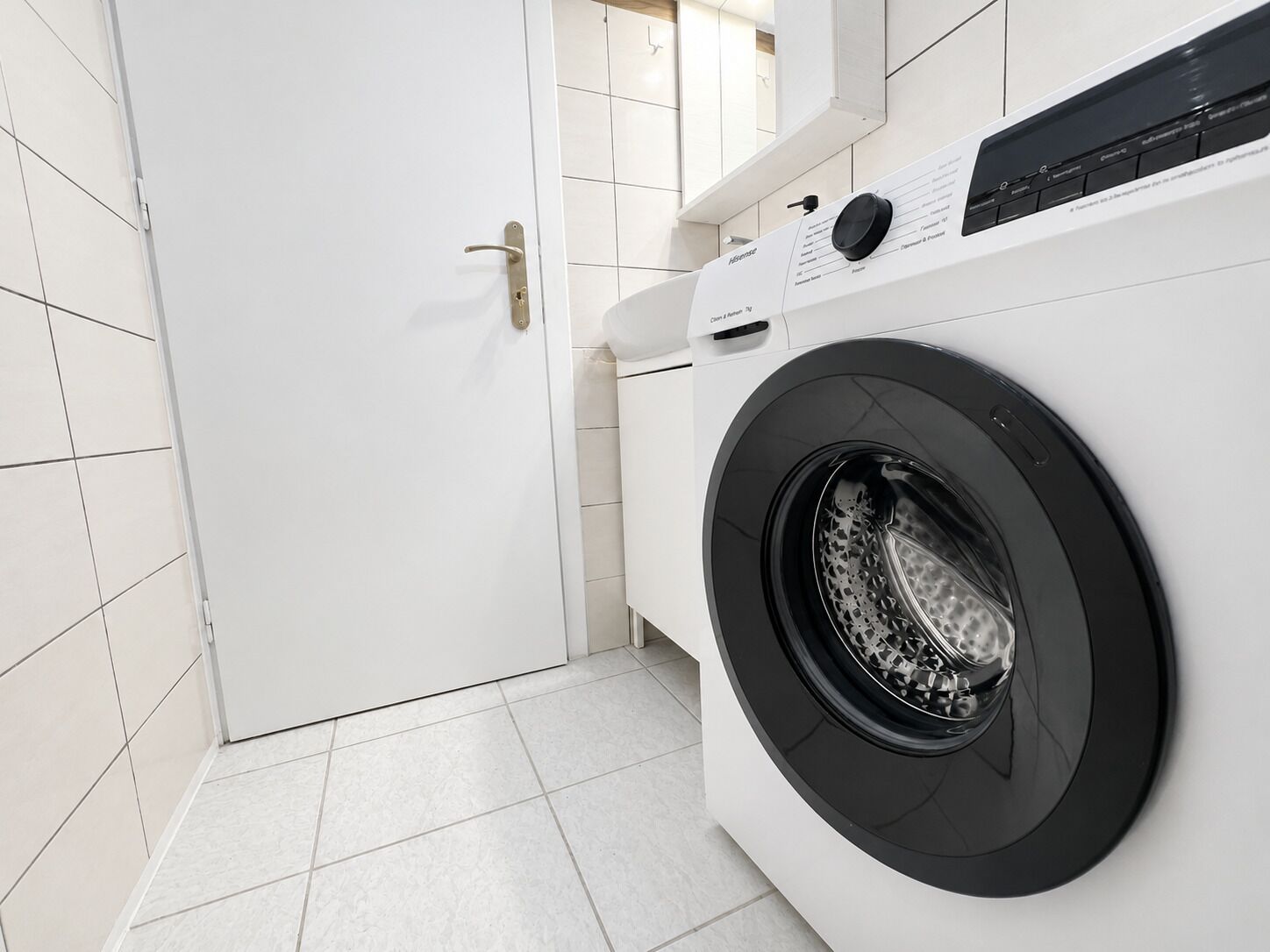 A modern laundry room with a front-loading washing machine, white tiled walls, and a door.