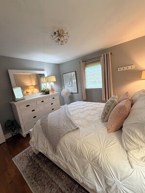 A bedroom with a large bed, a dresser, a mirror, a fan, and a window with curtains.