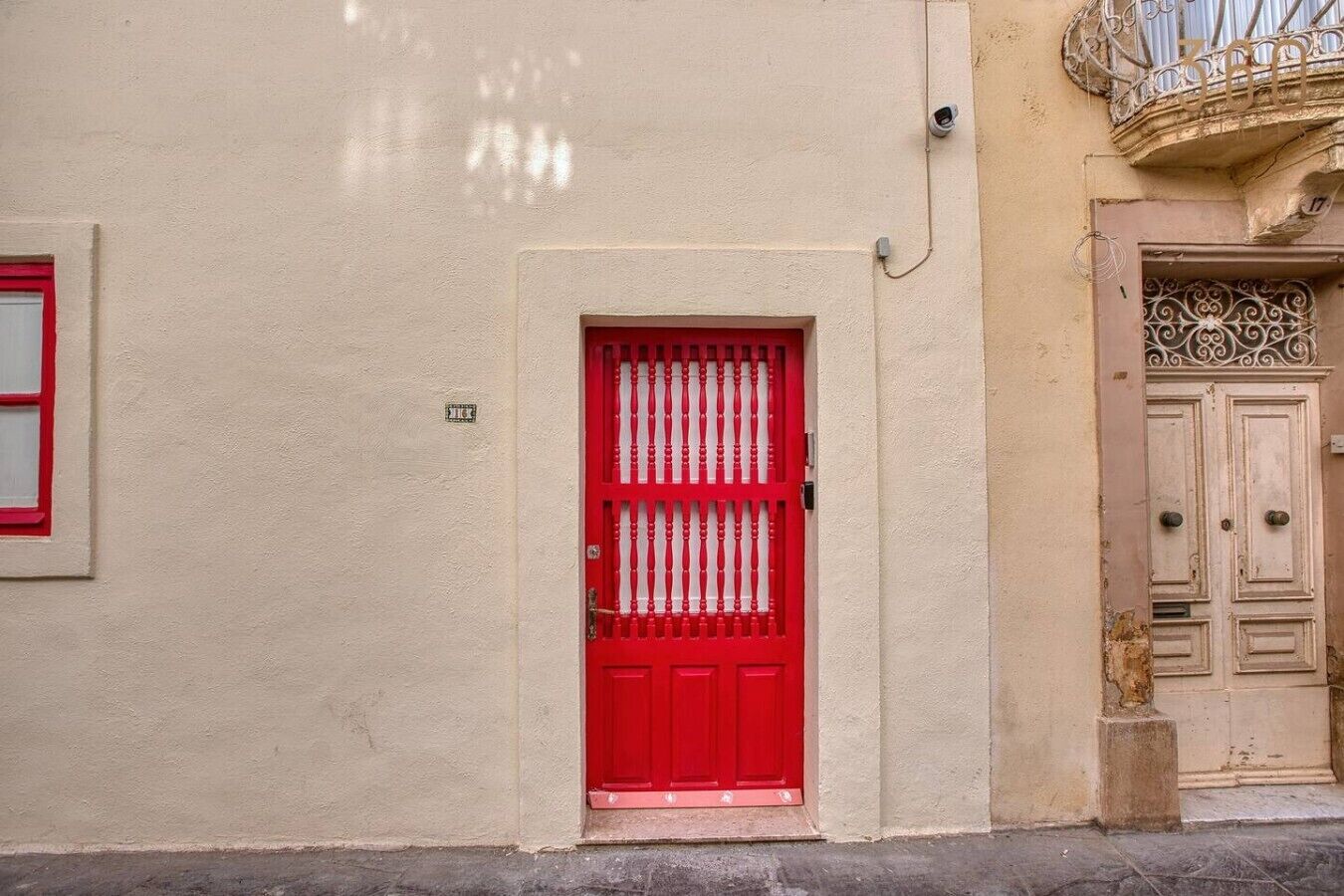 A red door with a metal grille, beige wall, and a balcony with ornate metalwork.