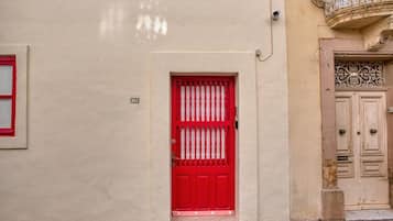 A red door with a metal grille, beige wall, and a balcony with ornate metalwork.