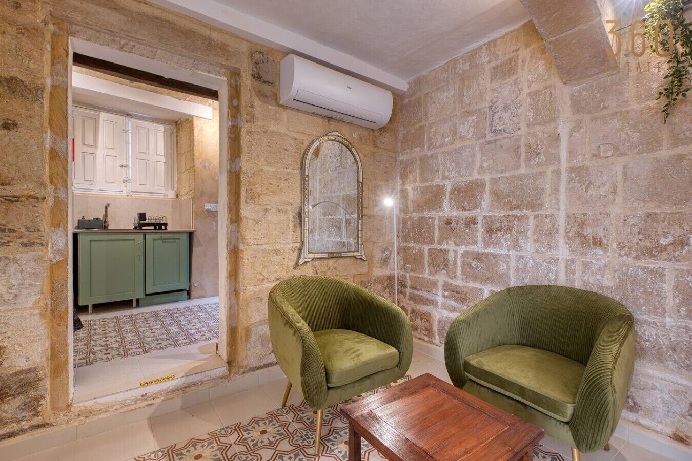 A room with stone walls, a mirror, two green chairs, a wooden table, and a kitchen area with green cabinets.