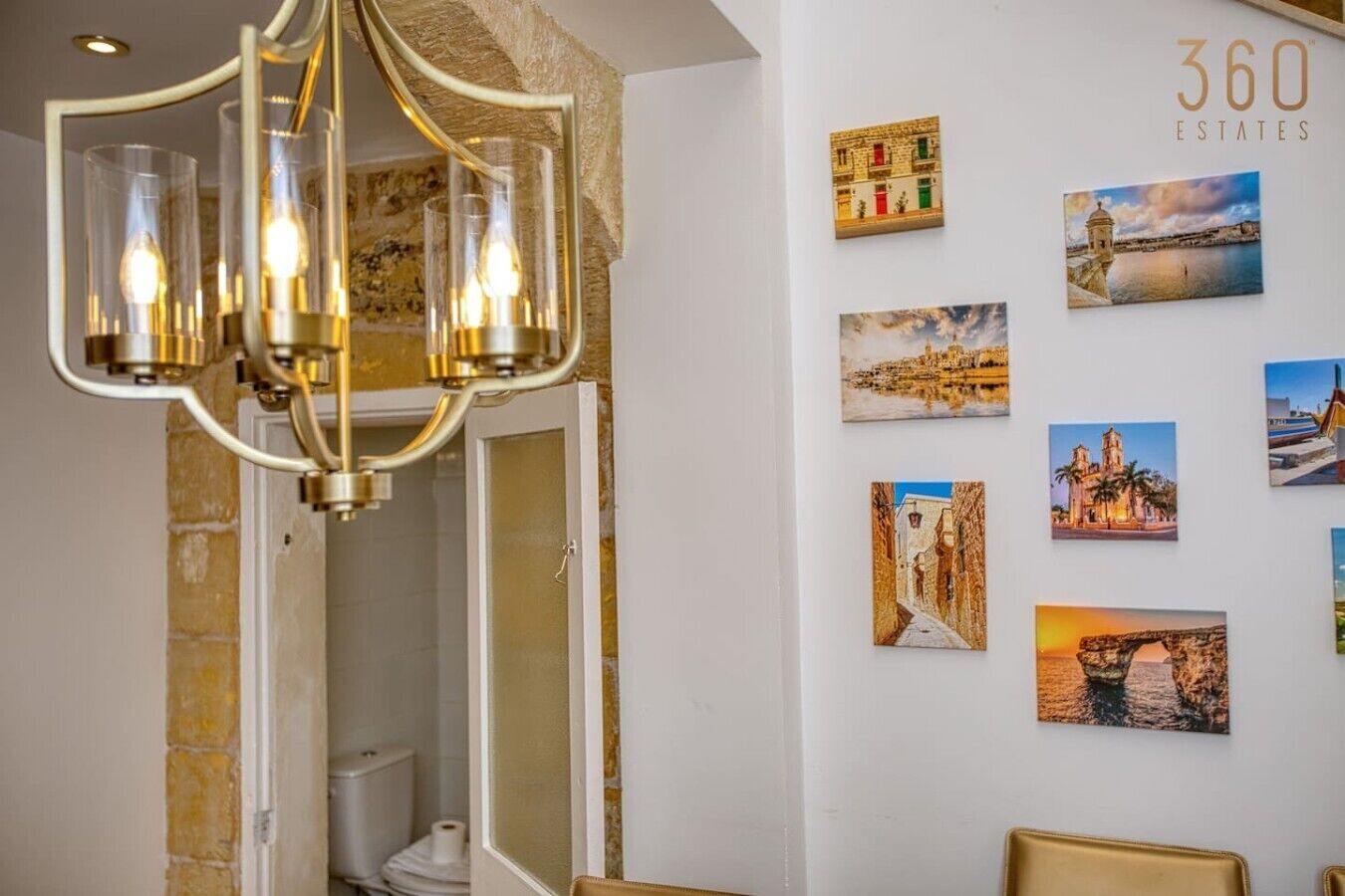 Et hotelbadeværelse med en moderne lysekrone, et hvidt toilet og en væg udsmykket med forskellige fotografier.