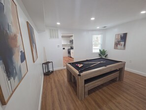 A room with a pool table, wooden flooring, white walls, and a kitchen area in the background. - Tidemark Row | Stylish Coastal Getaway (Atlantic City)