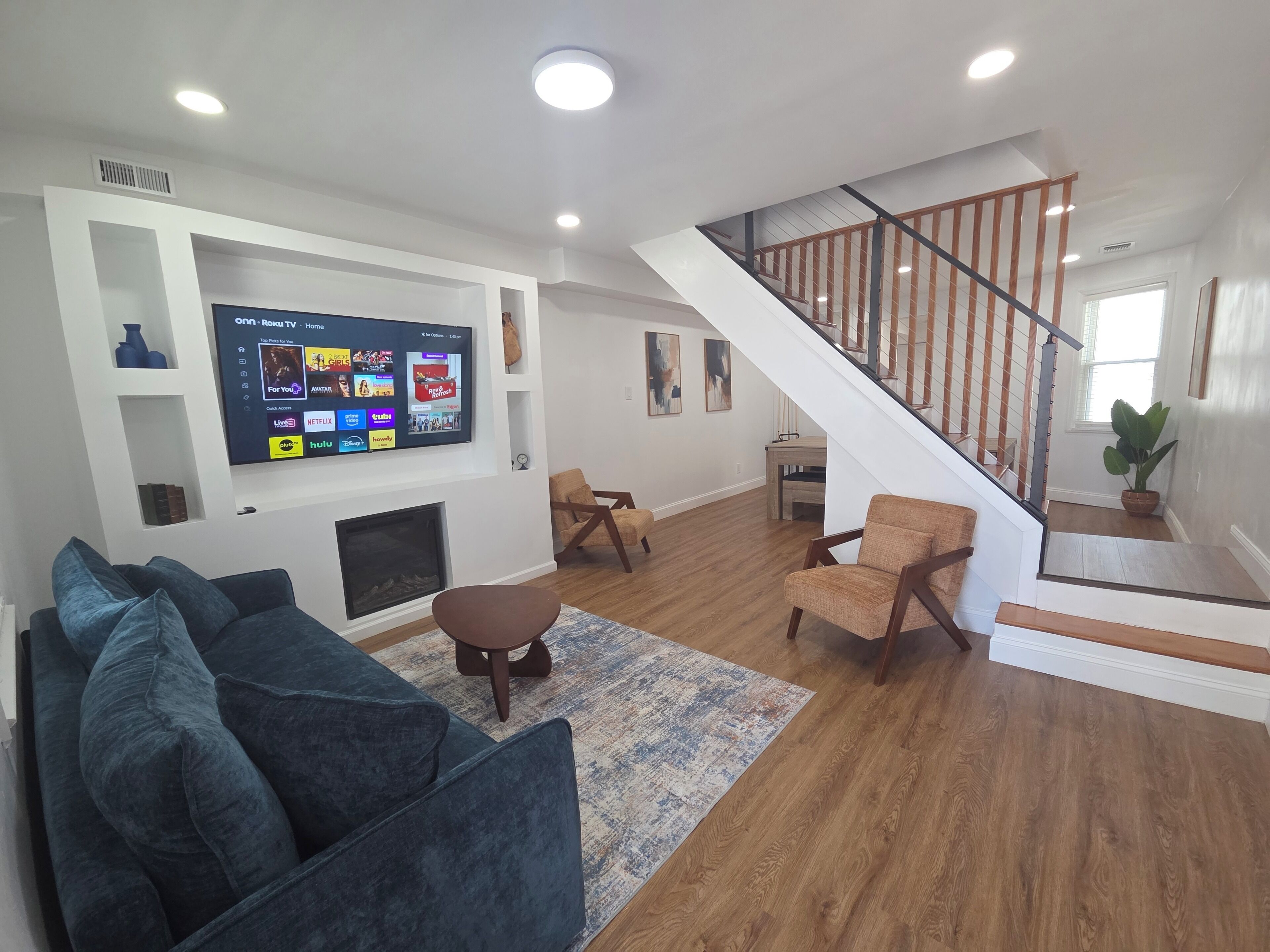 Un salon avec un téléviseur, un canapé, des fauteuils, une cheminée et un escalier.