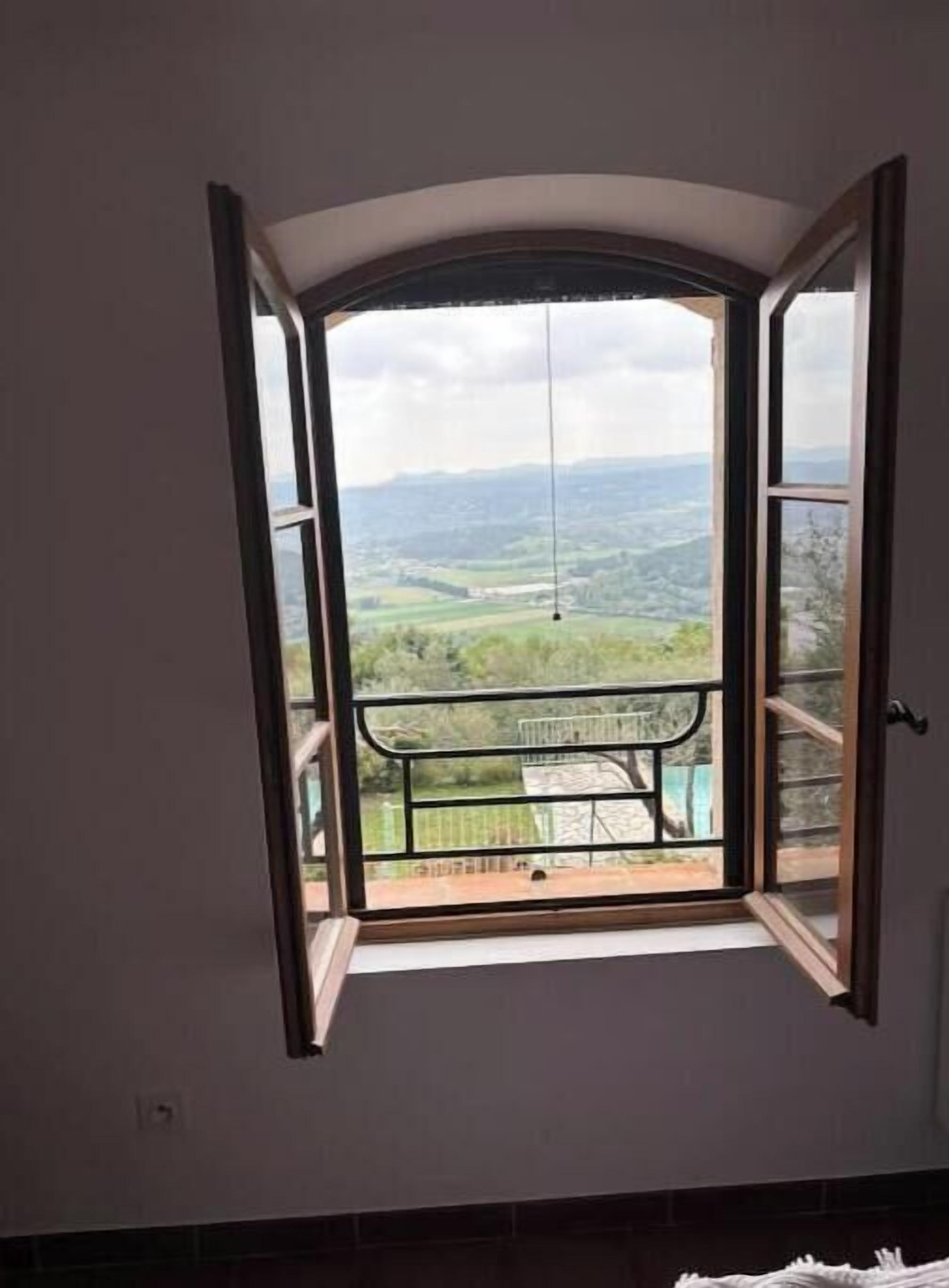 A room with a large window overlooking a balcony with a view of a green landscape and distant hills.