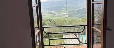 A room with a large window overlooking a balcony with a view of a green landscape and distant hills.