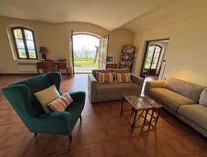 A spacious living room with a sofa, armchair, and a coffee table. There are two windows providing natural light and a view of the outdoors.