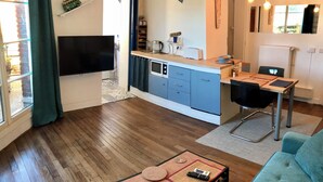 A modern living room with a sofa, a coffee table, a dining area with a table and chairs, a kitchen with blue cabinets, and a TV mounted on the wall. - Bright studio apartment with a small terrace (Viroflay)
