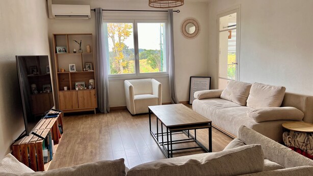 Un salon comprenant un canapé, une table basse, un fauteuil, une bibliothèque en bois et une fenêtre donnant sur des arbres.