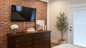 Una habitación con pared de ladrillo, un televisor de pantalla plana montado sobre un tocador de madera, una planta en maceta y una puerta con persianas.