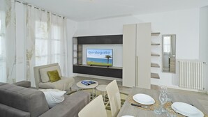 A modern living room with a flat-screen TV, a sofa, a dining table set for four, and a large window with curtains. - Corte Fasanella 3 in Toscolano-maderno (Toscolano Maderno)