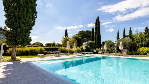 A rectangular swimming pool with a tiled deck, surrounded by lounge chairs and umbrellas, with tall trees and a clear sky in the background. - Corte Fasanella 3 in Toscolano-maderno (Toscolano Maderno)