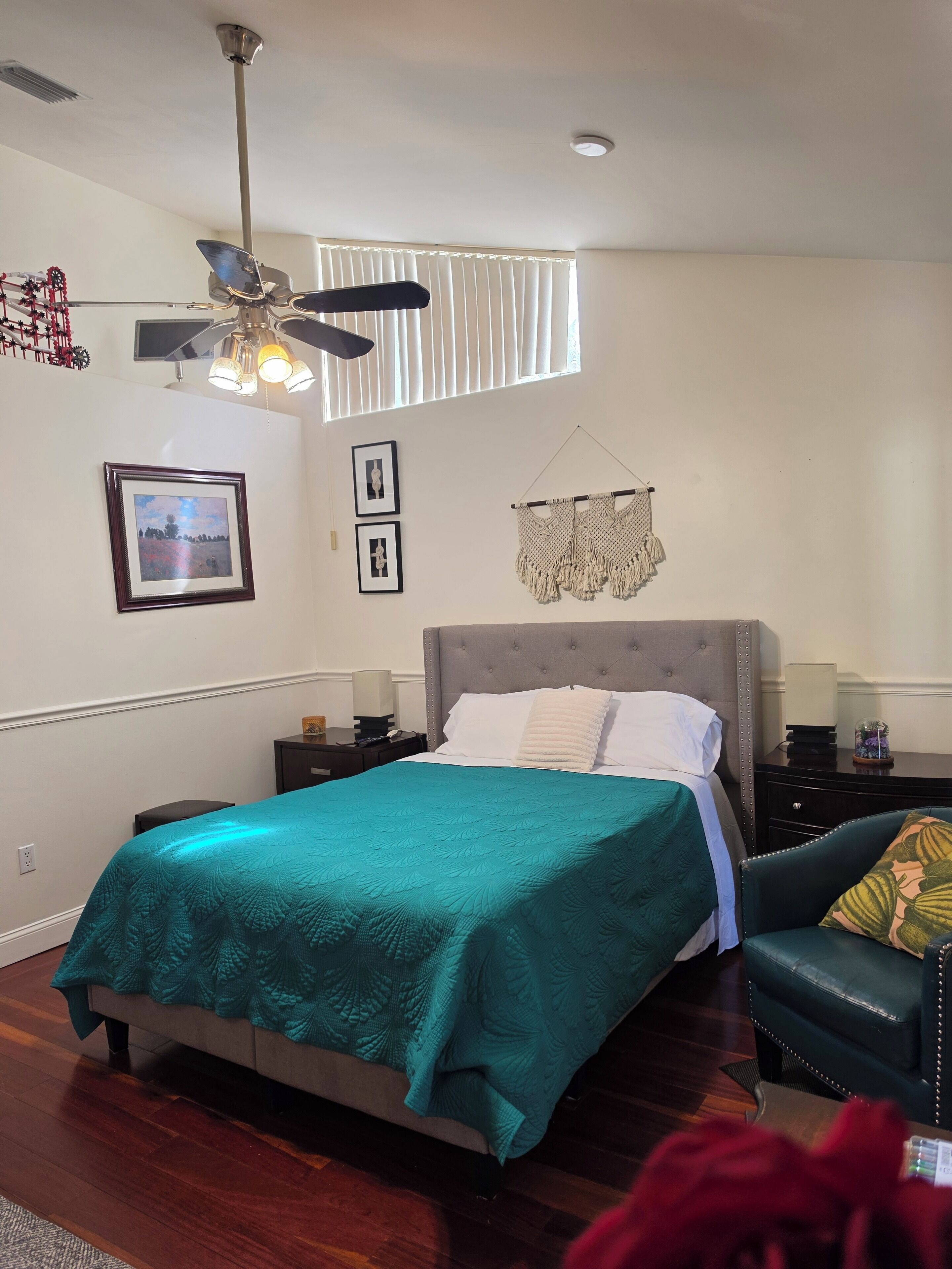 A bedroom with a bed, a ceiling fan, a nightstand, a chair, and a wall-mounted macrame piece.