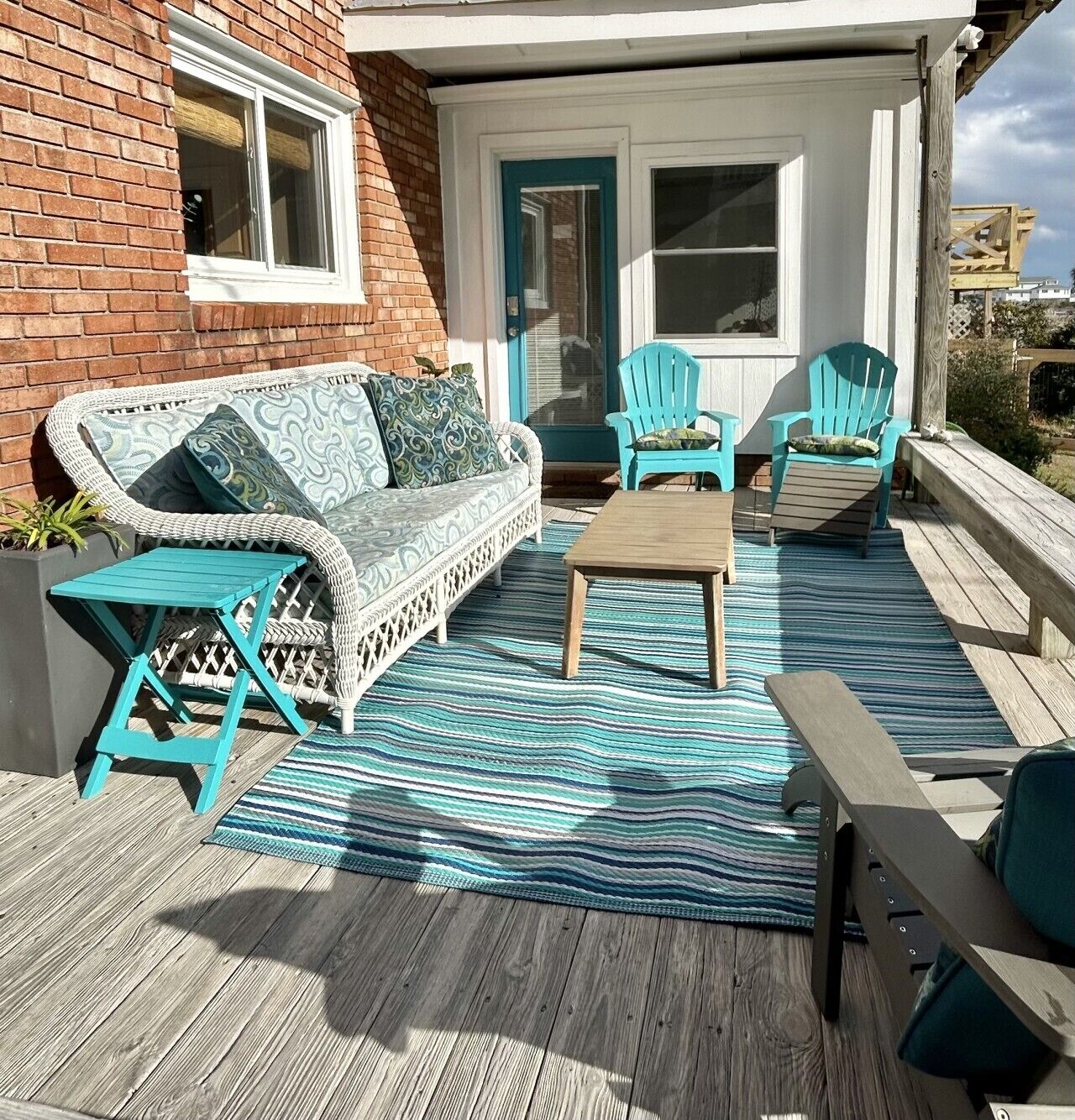 A covered outdoor seating area with a wicker sofa, two Adirondack chairs, a wooden table, and a striped rug.