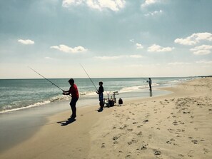 A beach with people fishing, a cart with fishing gear, and footprints in the sand. - The Beach Club | Classic AB Cottage by The Circle (Atlantic Beach)