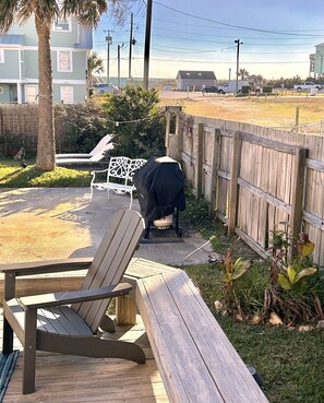 Terraza de madera con una silla de descanso, una parrilla cubierta y un patio cercado con vista al océano.