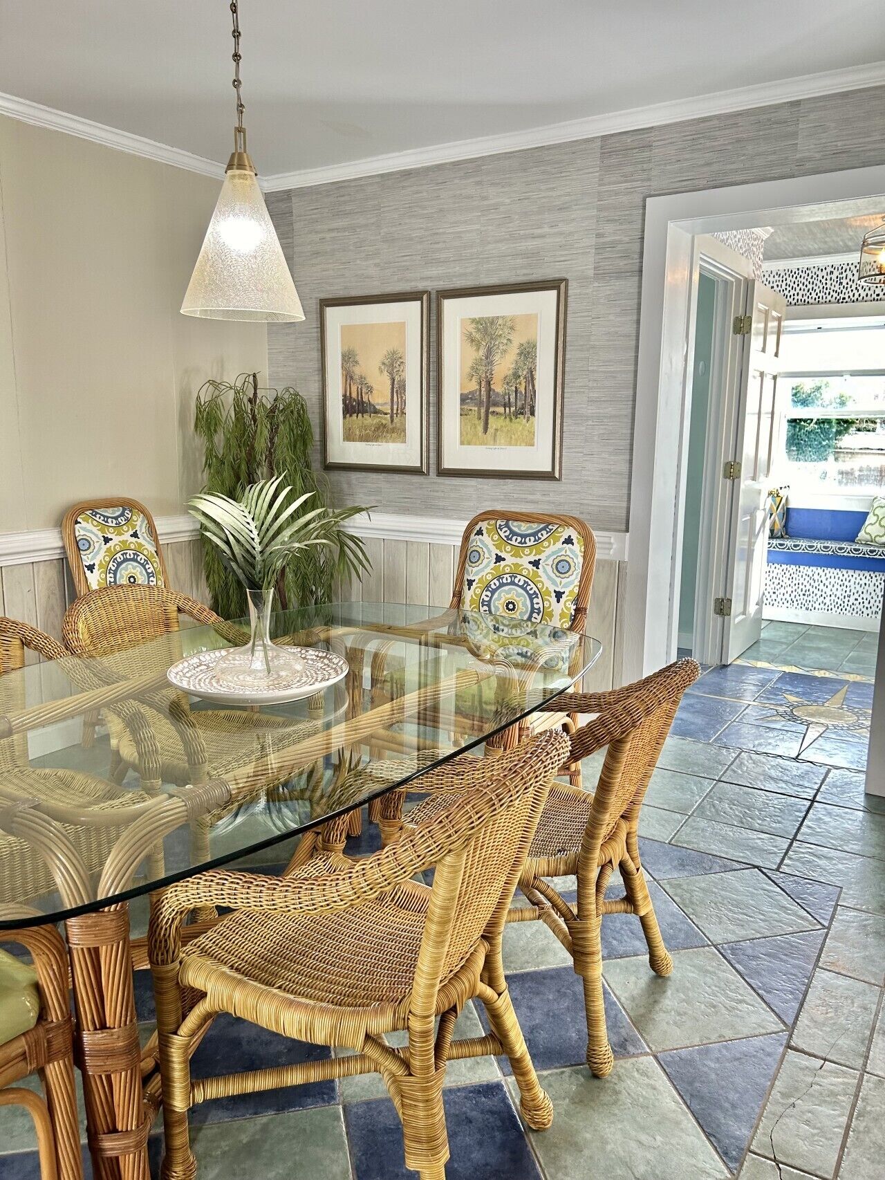 A dining area with a glass-top table, wicker chairs, a hanging light fixture, framed artwork on the wall, and a potted plant.