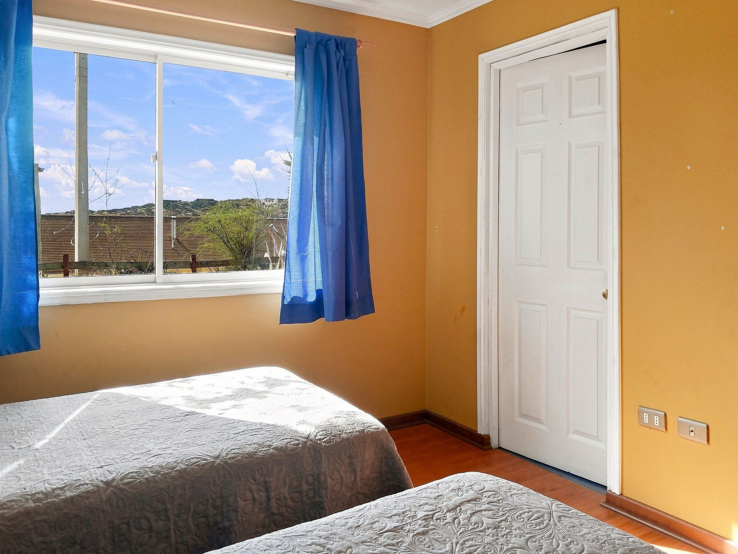 Une chambre avec deux lits, une fenêtre avec des rideaux, une porte blanche et une vue sur l’extérieur.