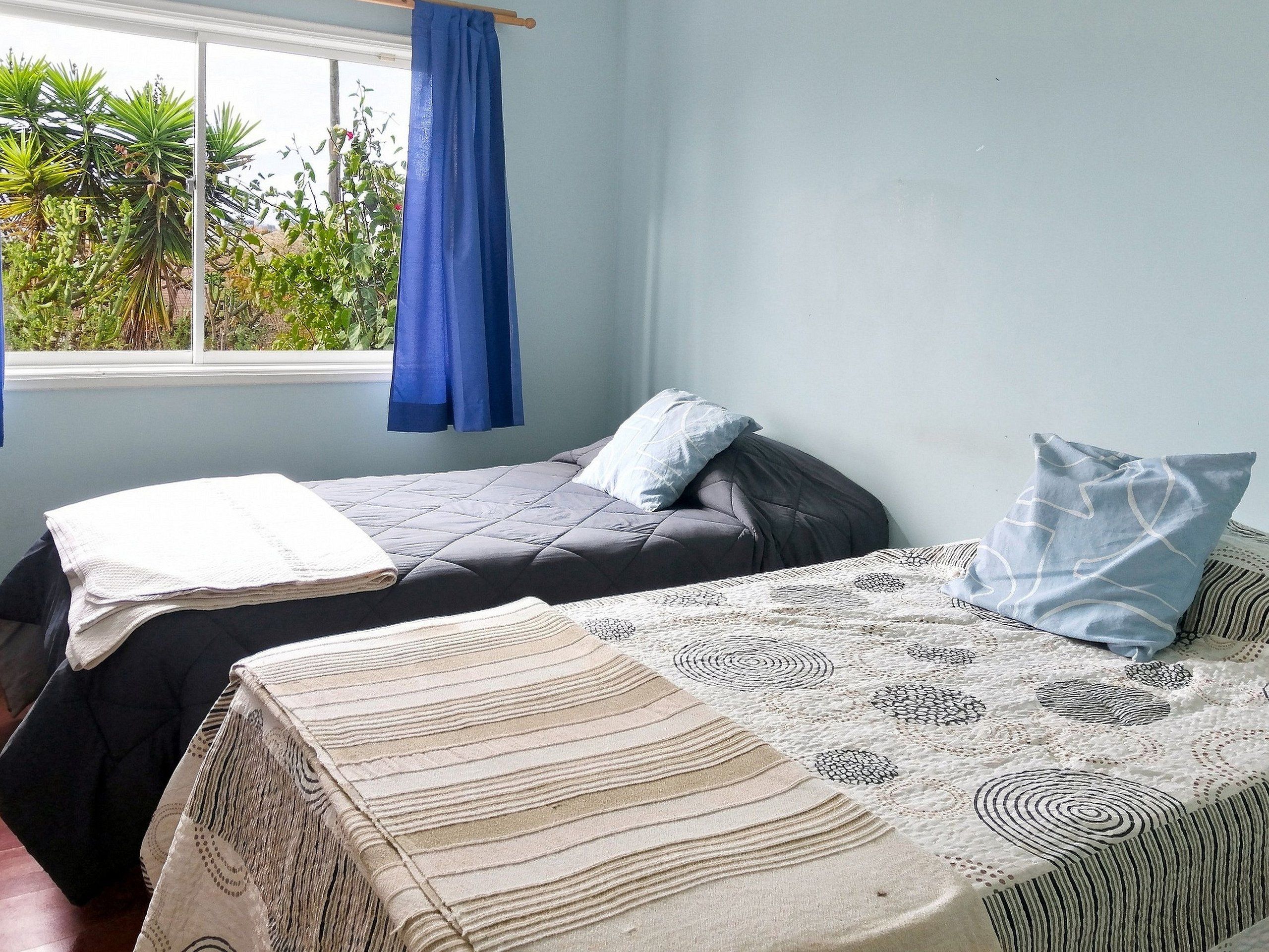 Ein Zimmer mit zwei Betten, eines mit einer gemusterten Tagesdecke und das andere mit einer schlichten. Ein Fenster mit blauen Vorhängen bietet Blick auf Grünflächen.