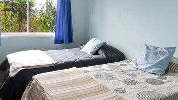 A room with two beds, one with a patterned bedspread and the other with a plain one. There is a window with blue curtains, and a view of greenery outside.