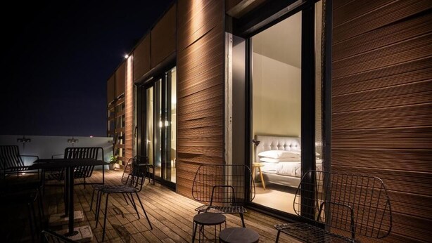 A modern building exterior with a wooden facade, large glass doors leading to a bedroom with a bed and a nightstand, and outdoor seating with metal chairs and tables. - Aurum ApartHotels Città Studi (Milano)