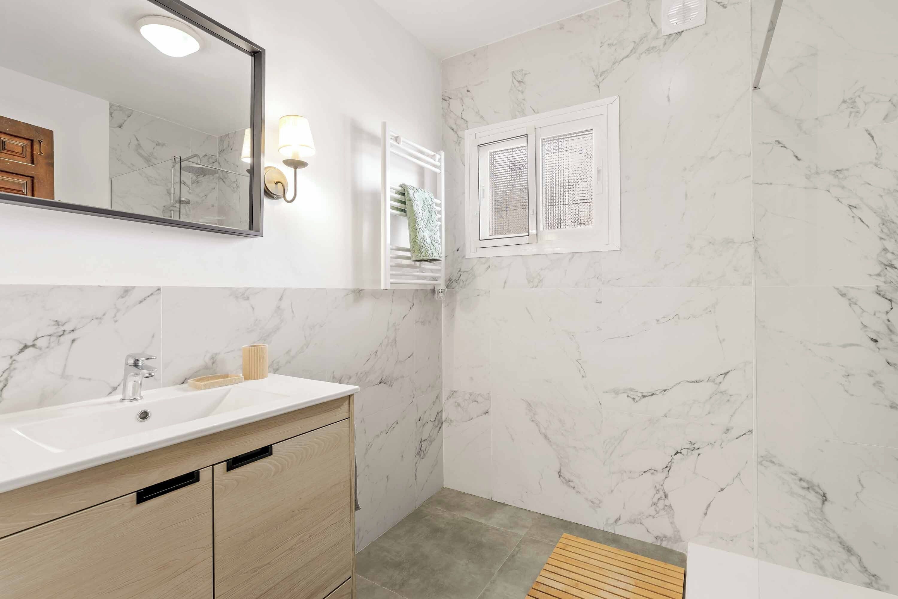 A bathroom with a marble wall, a sink with a mirror, a towel rack, and a window with a view.