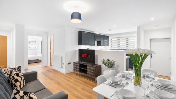 A modern living room with a sofa, a dining table set for four, a TV displaying 'NETFLIX,' and a kitchen area with cabinets and a window. - Urban Shore | By My Getaways (Brighton)
