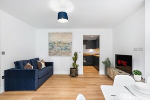 Un salon moderne avec un canapé, un téléviseur affichant le logo « NETFLIX », et une cuisine en arrière-plan.