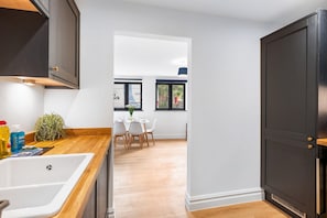 Eine moderne Küche mit Holzarbeitsplatte, weißem Spülbecken und dunklen Schränken. Es gibt eine Essecke mit Tisch und Stühlen sowie einen Blick ins Freie durch ein Fenster.