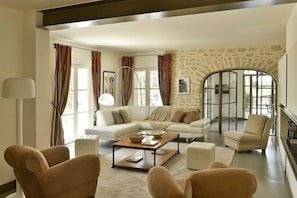 A spacious living room with a large sofa, a coffee table, and two armchairs. There is a stone wall and a large window with curtains. - Mas Saint Jean - Domaine de Malaga - New Porperty (Maussane-les-Alpilles)