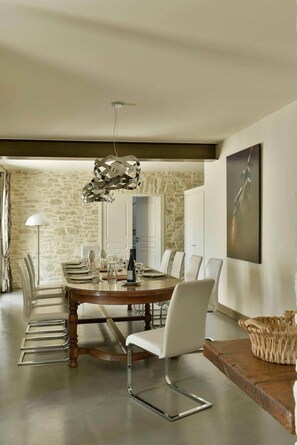 A dining room with a long table, white chairs, a modern pendant light, and a stone wall. - Mas Saint Jean - Domaine de Malaga - New Porperty (Maussane-les-Alpilles)