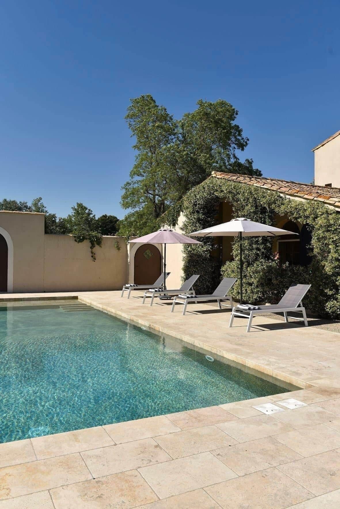 A swimming pool with clear water, surrounded by lounge chairs and umbrellas, and a building with a tiled roof and greenery.