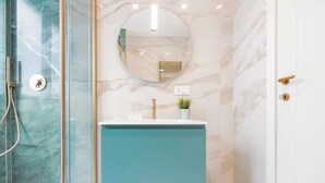 Une salle de bain dotée d’une douche à parois de verre, d’un mur en marbre, d’un miroir rond, d’un meuble-lavabo de couleur turquoise et d’une poignée de porte dorée.