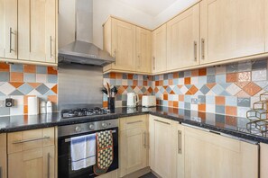 A kitchen with wooden cabinets, a gas stove, a range hood, and a tiled backsplash. - GuestReady - Charming Kensington Flat (London)