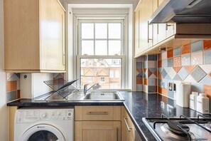 A kitchen with a washing machine, a sink, a stove, and a window with a view of buildings. - GuestReady - Charming Kensington Flat (London)