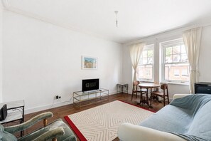 A room with a bed, a TV, a dining table with chairs, a small table with a chair, a window with curtains, and a picture on the wall. - GuestReady - Charming Kensington Flat (London)