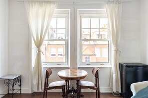 A room with two windows, a round table with chairs, a small metal table, and a black appliance. - GuestReady - Charming Kensington Flat (London)