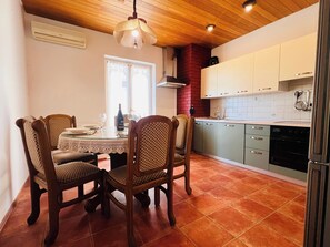 A dining area with a table set for four, wooden chairs, a chandelier, and a kitchen with white cabinets and a red brick wall. - A6 - Spacious apt w Pool, Terrace and sea View (Dugi Rat)